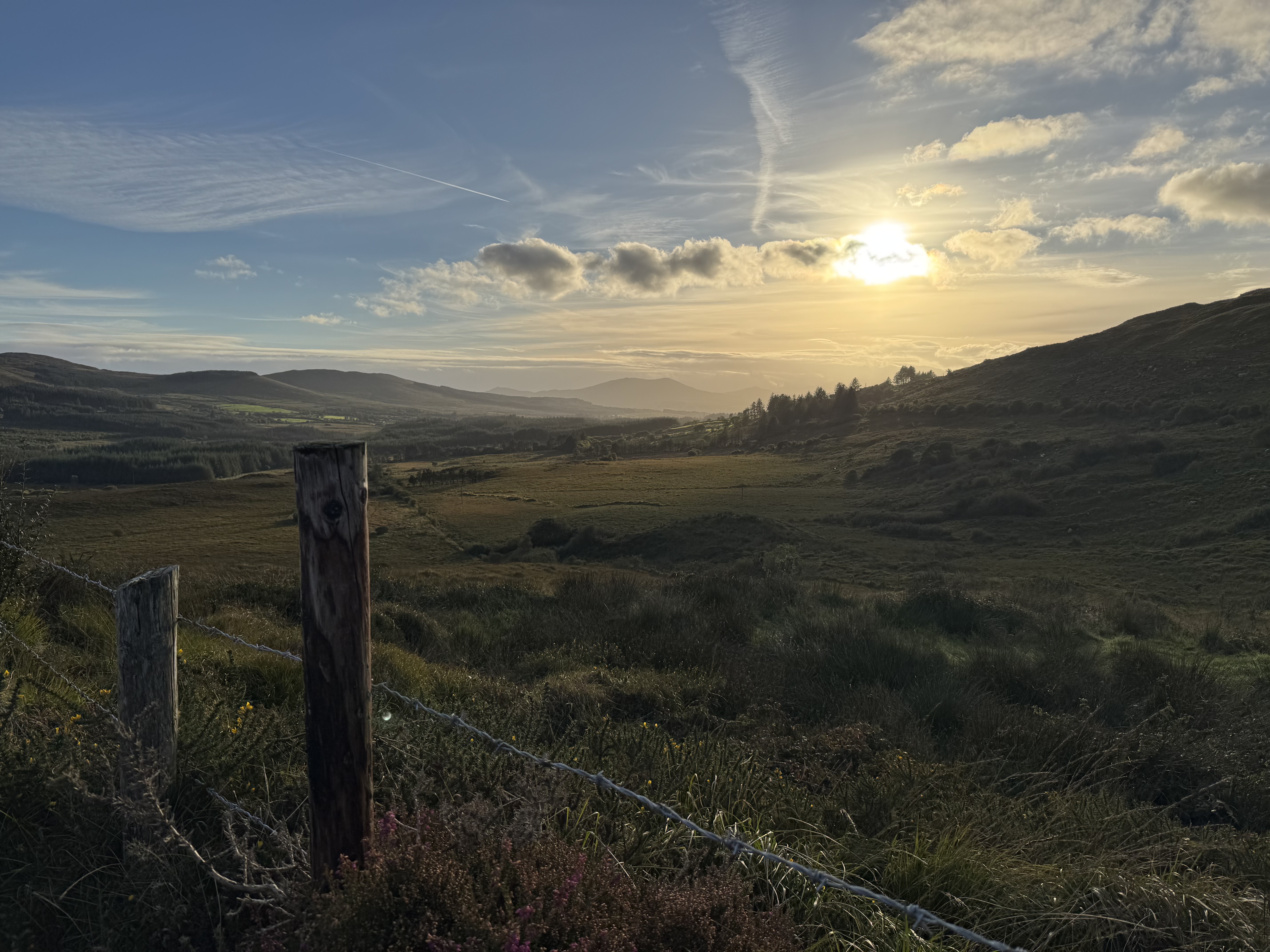 Scenic view of rolling hills and valleys at sunset, with a wooden fence post in the foreground and wispy clouds in the sky.