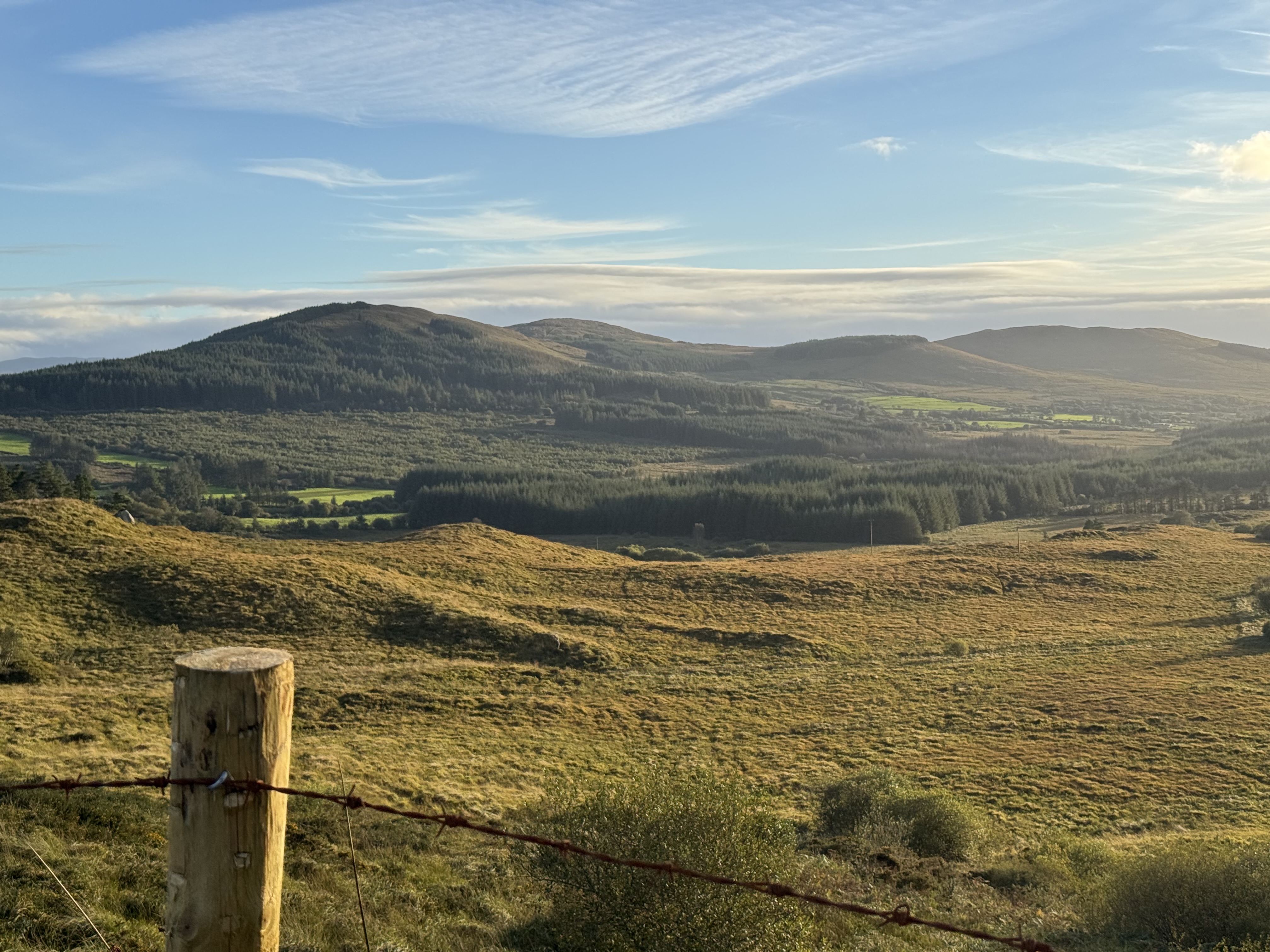 A scenic view of rolling hills and valleys under a blue sky with scattered clouds, featuring lush greenery and a wooden post with barbed wire in the foreground.